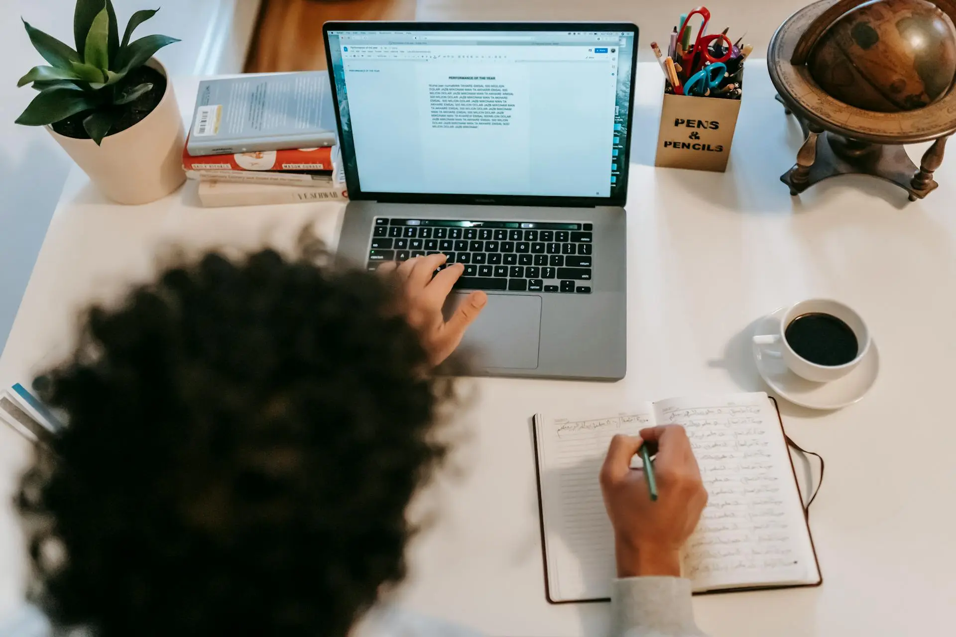 Overhead shot of an author working on a laptop while writing in a notebook